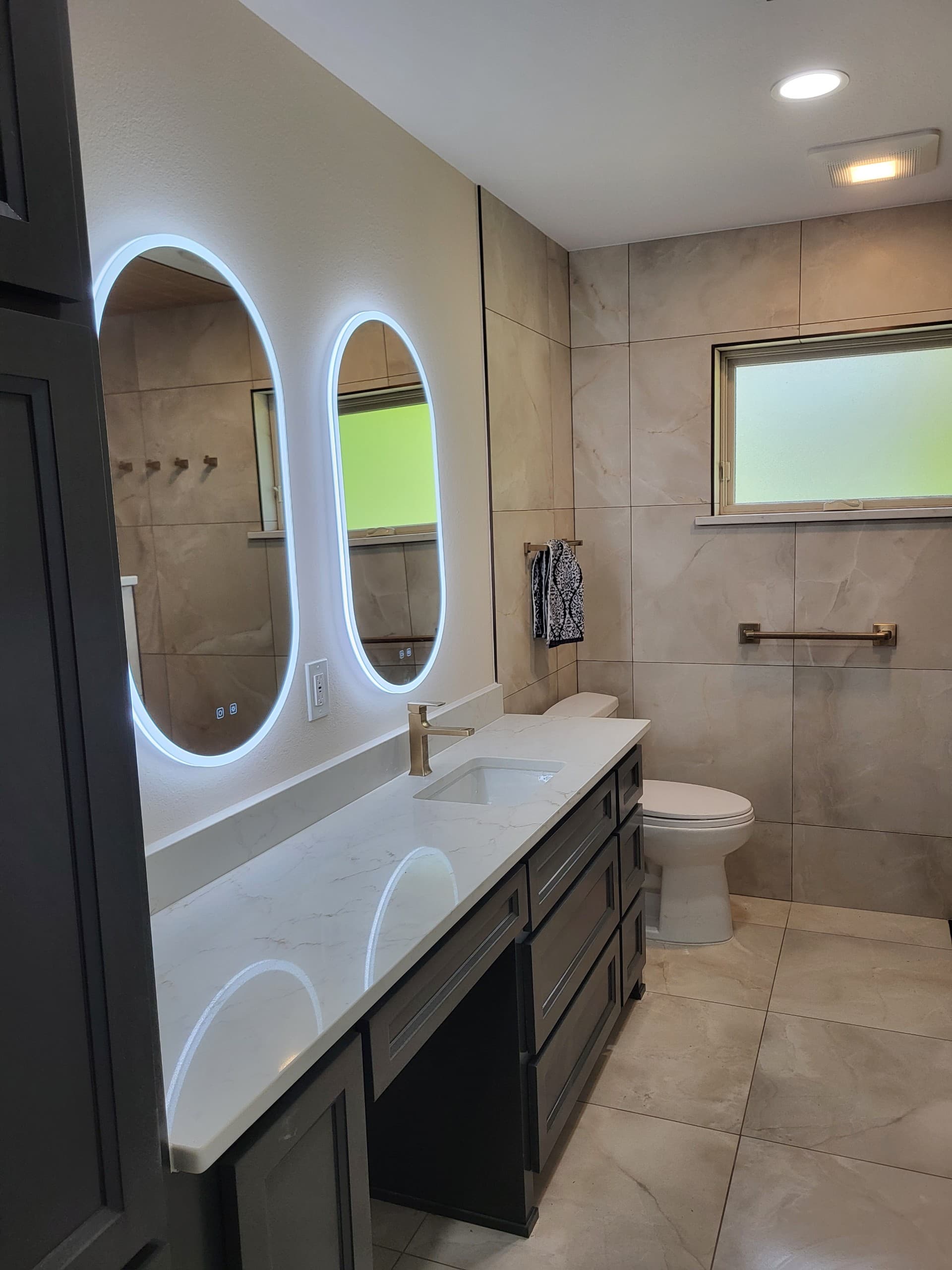 Modern bathroom featuring two illuminated oval mirrors above a white marble vanity with gold fixtures.