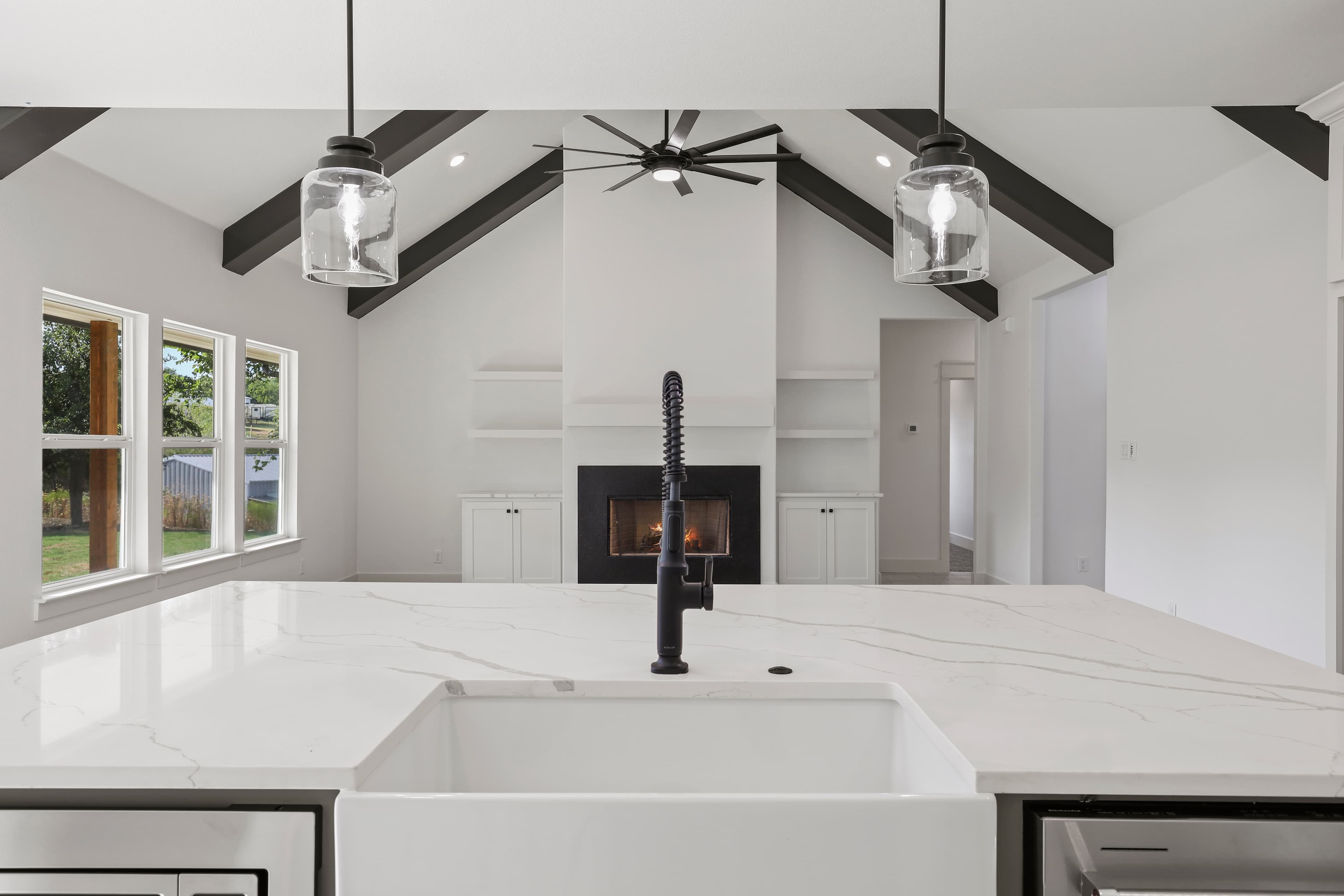 White marble kitchen island with farmhouse sink facing a vaulted living room with dark beams.