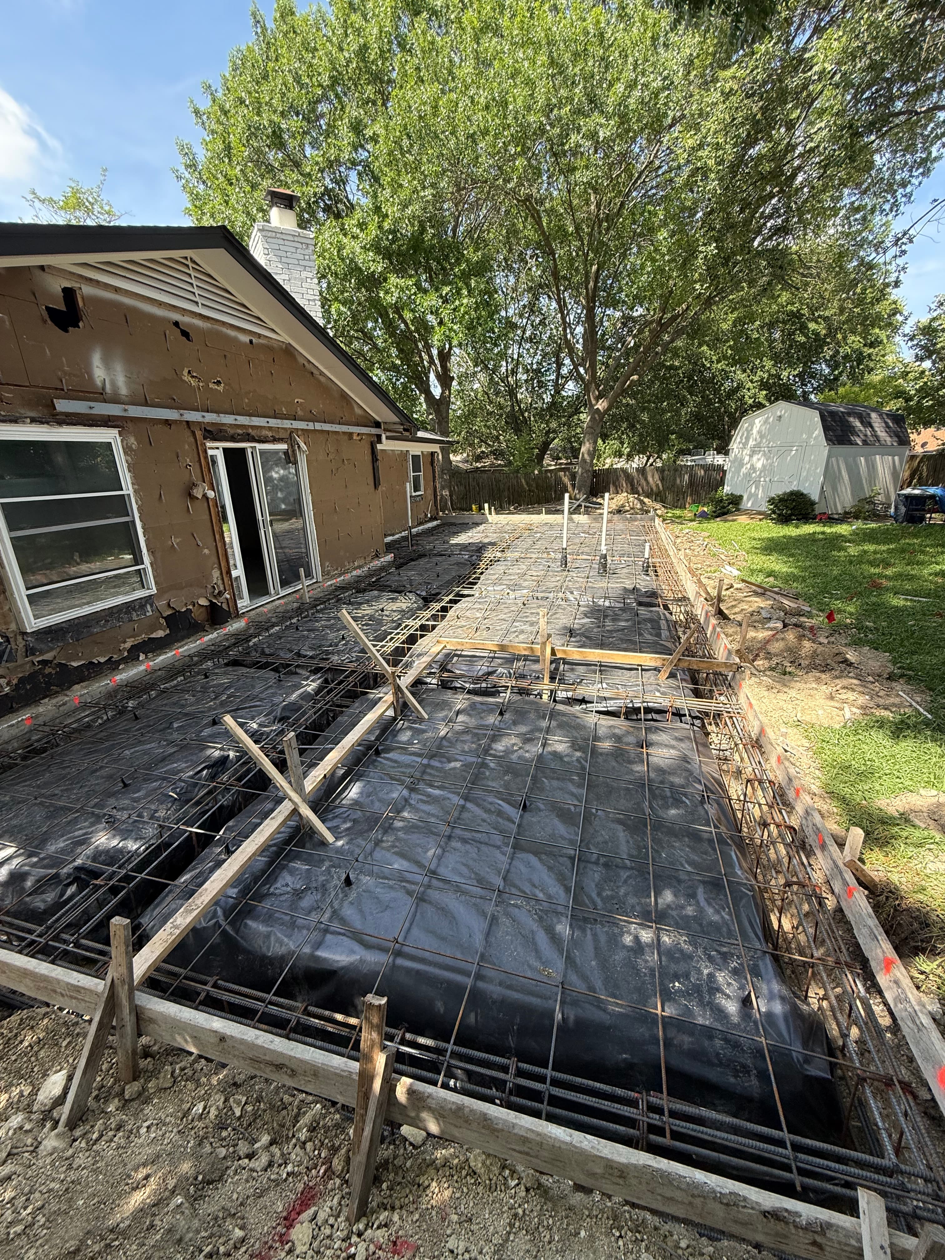 House foundation under construction with rebar grid over black plastic and wooden forms.