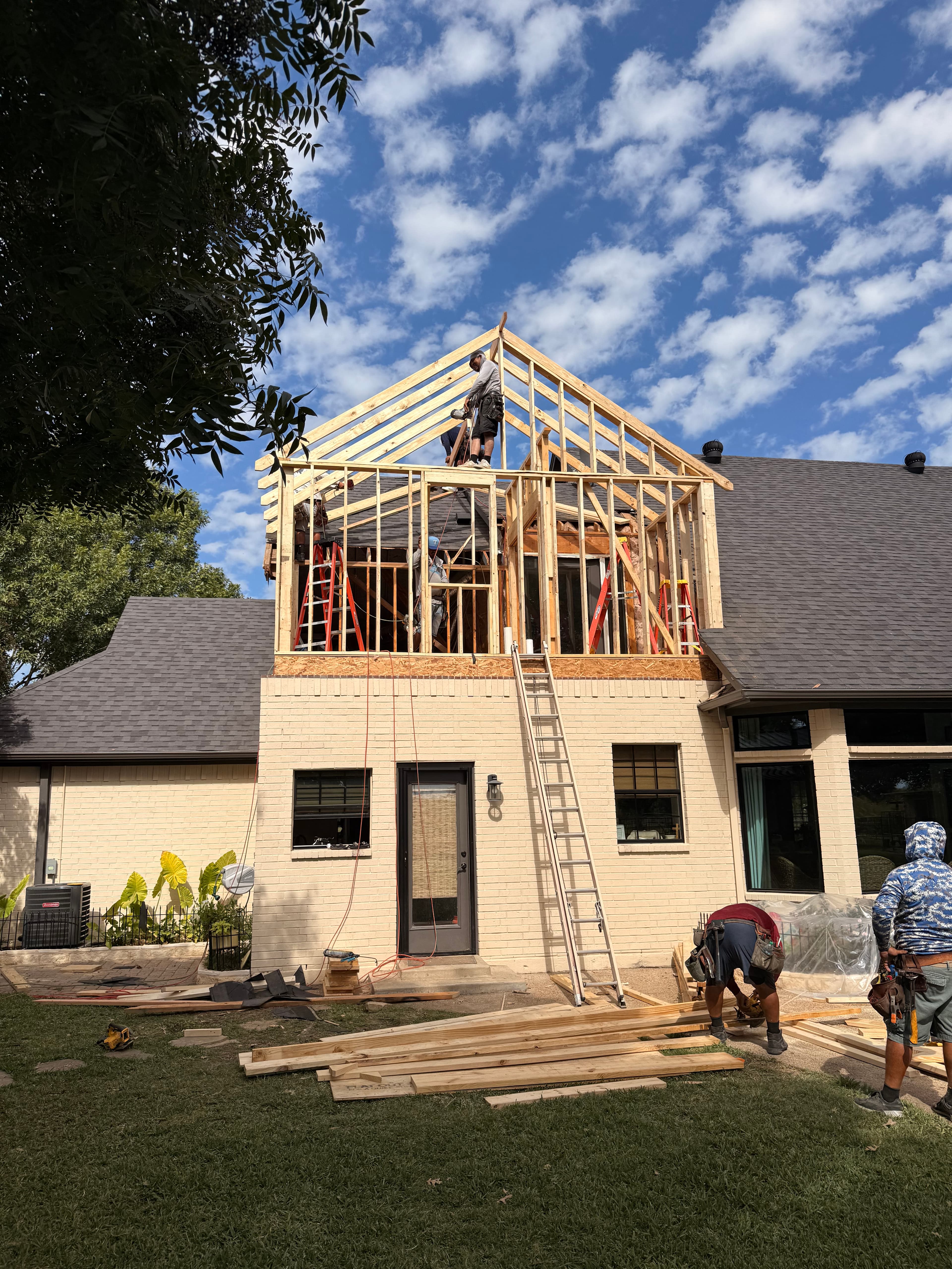 Construction workers framing a new wooden roof addition on a brick house under blue skies.