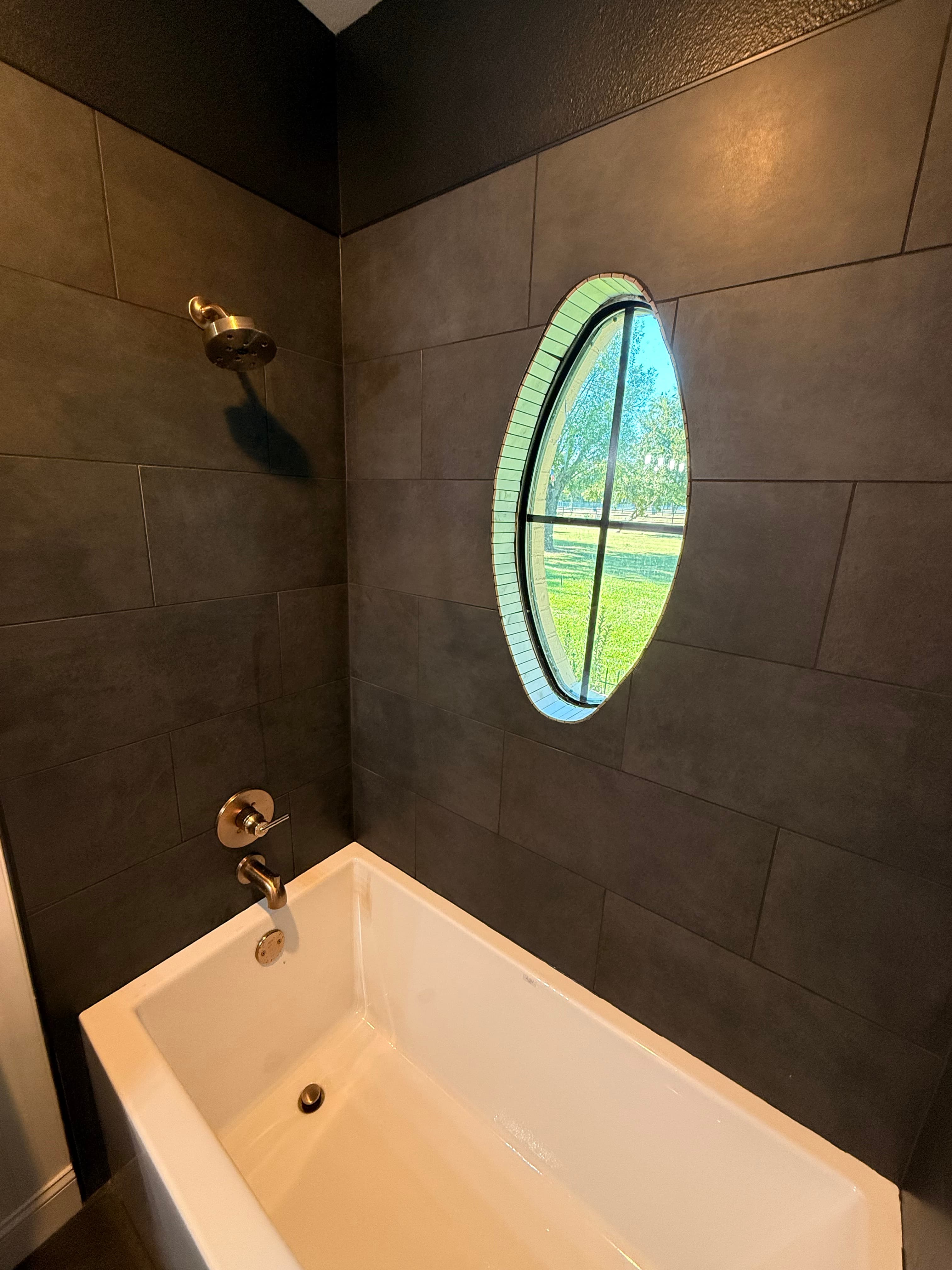 Modern bathroom featuring dark grey tiles, brass fixtures, and an oval window overlooking greenery.