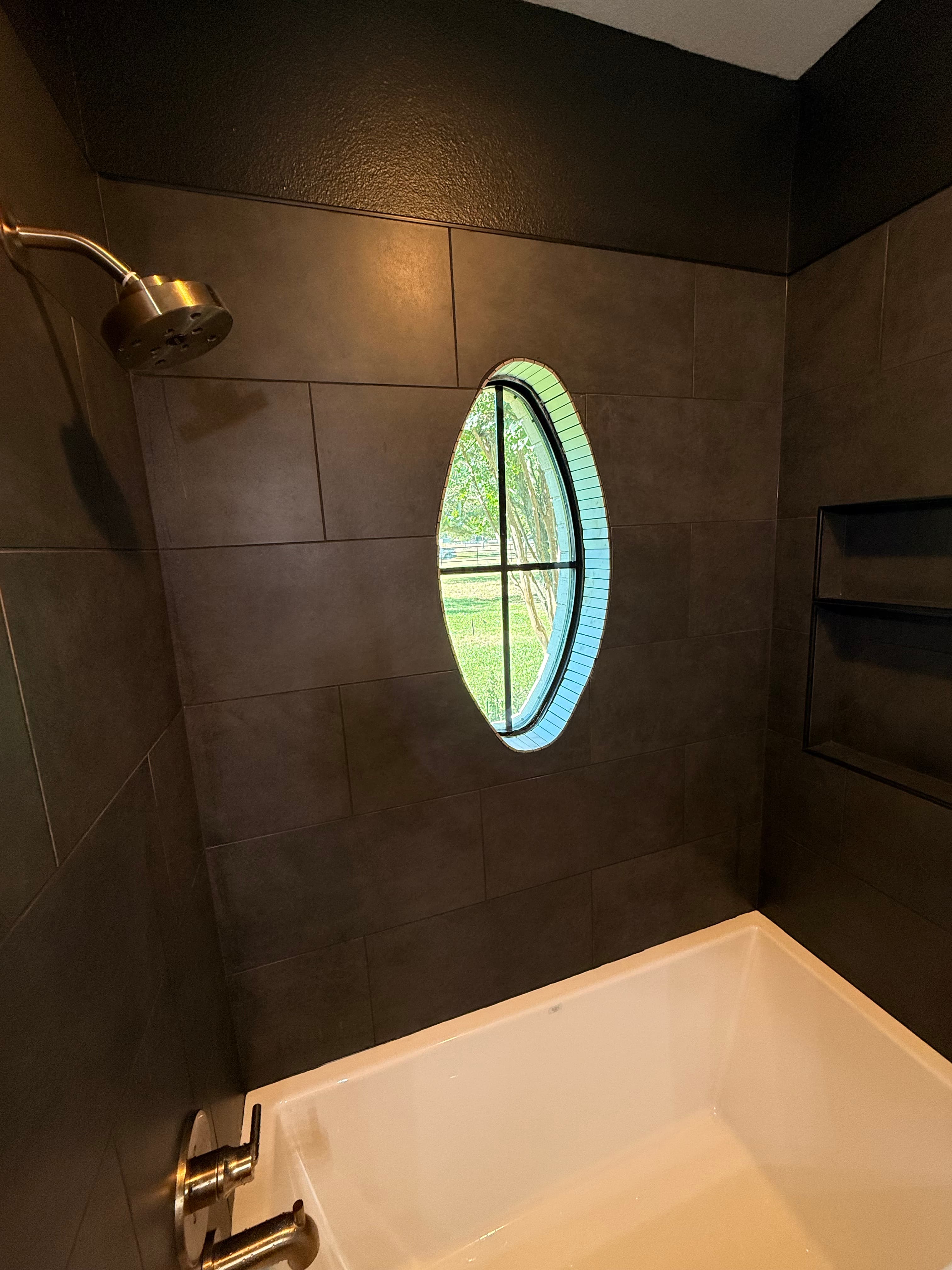 Modern dark-tiled bathroom with an oval window showing trees, a showerhead, and a bathtub.