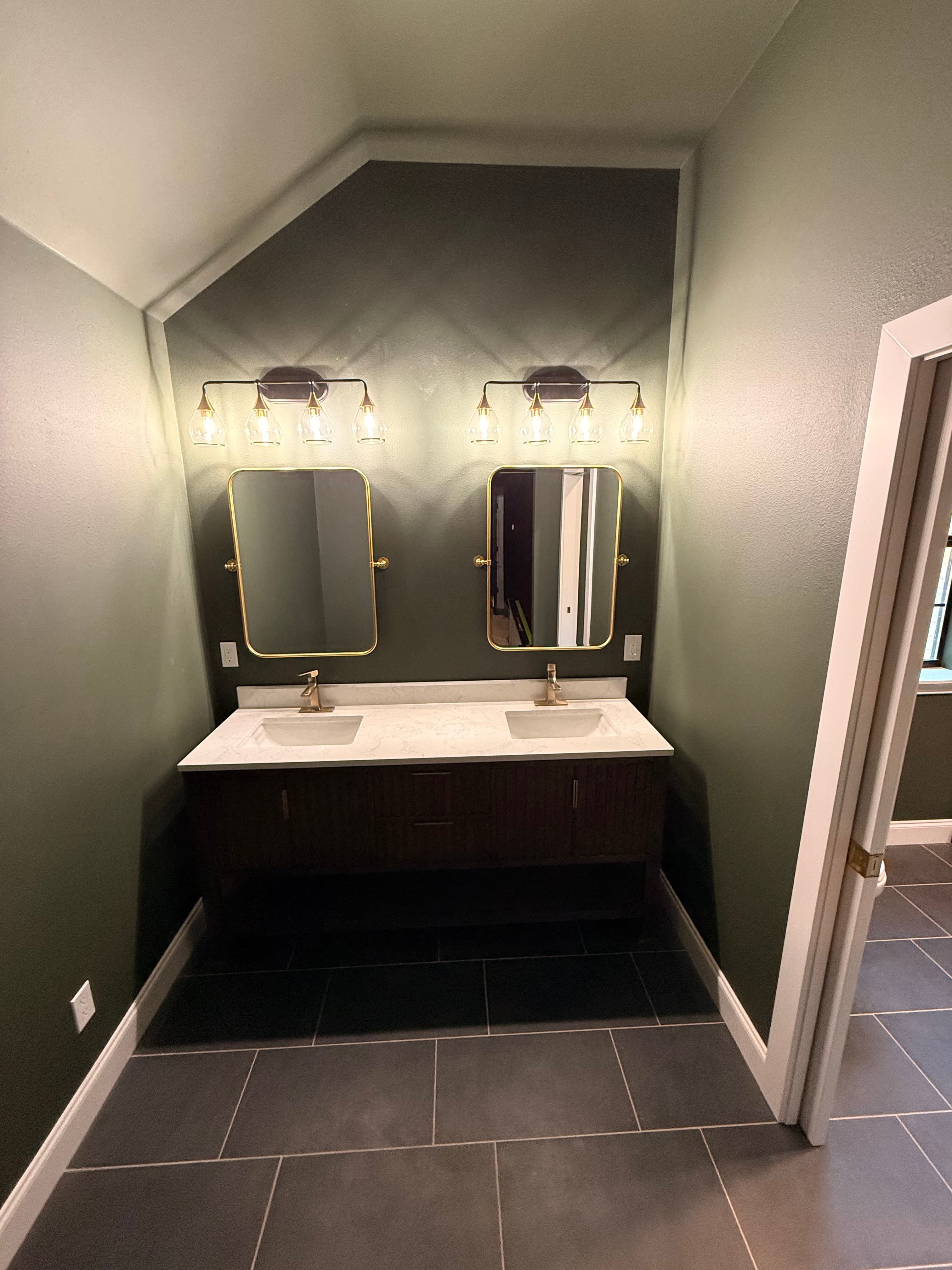 Dark green bathroom wall featuring a double vanity, gold-framed mirrors, and matching gold lighting.
