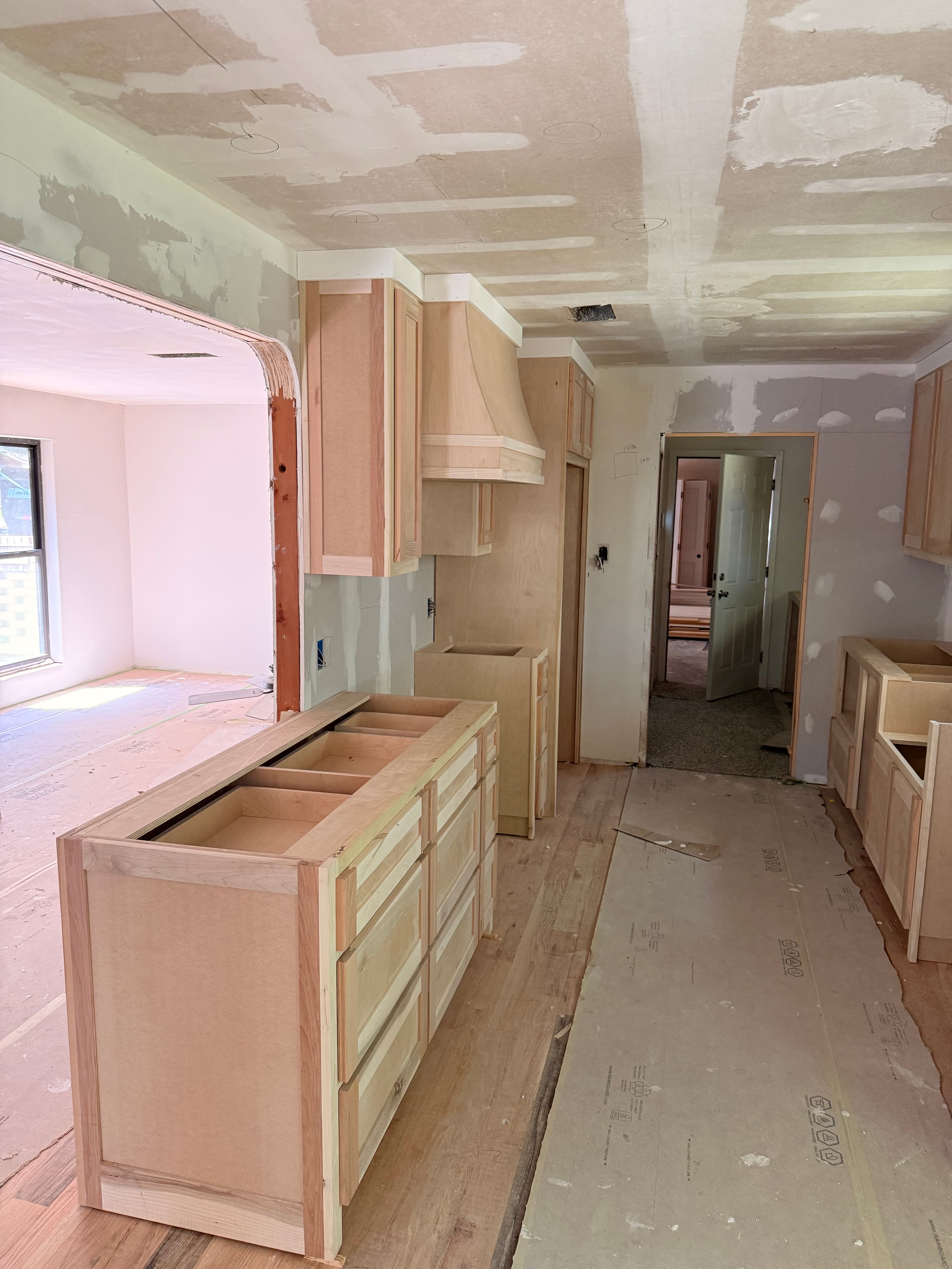 Kitchen under renovation with unfinished wood cabinets, mudded drywall walls, and protective floor covering.