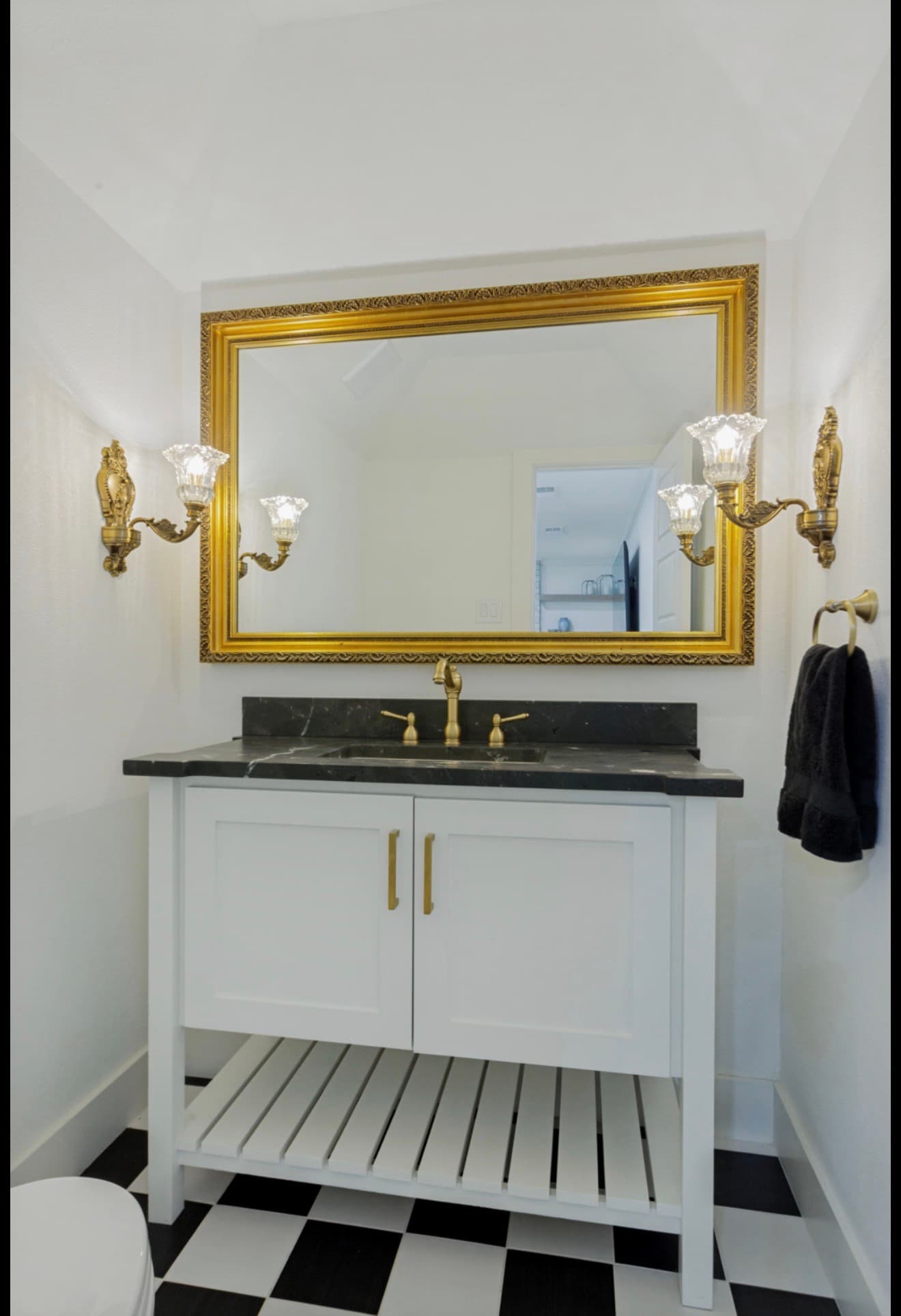 White vanity with black marble top, gold mirror, ornate sconces, and a checkered tile floor.