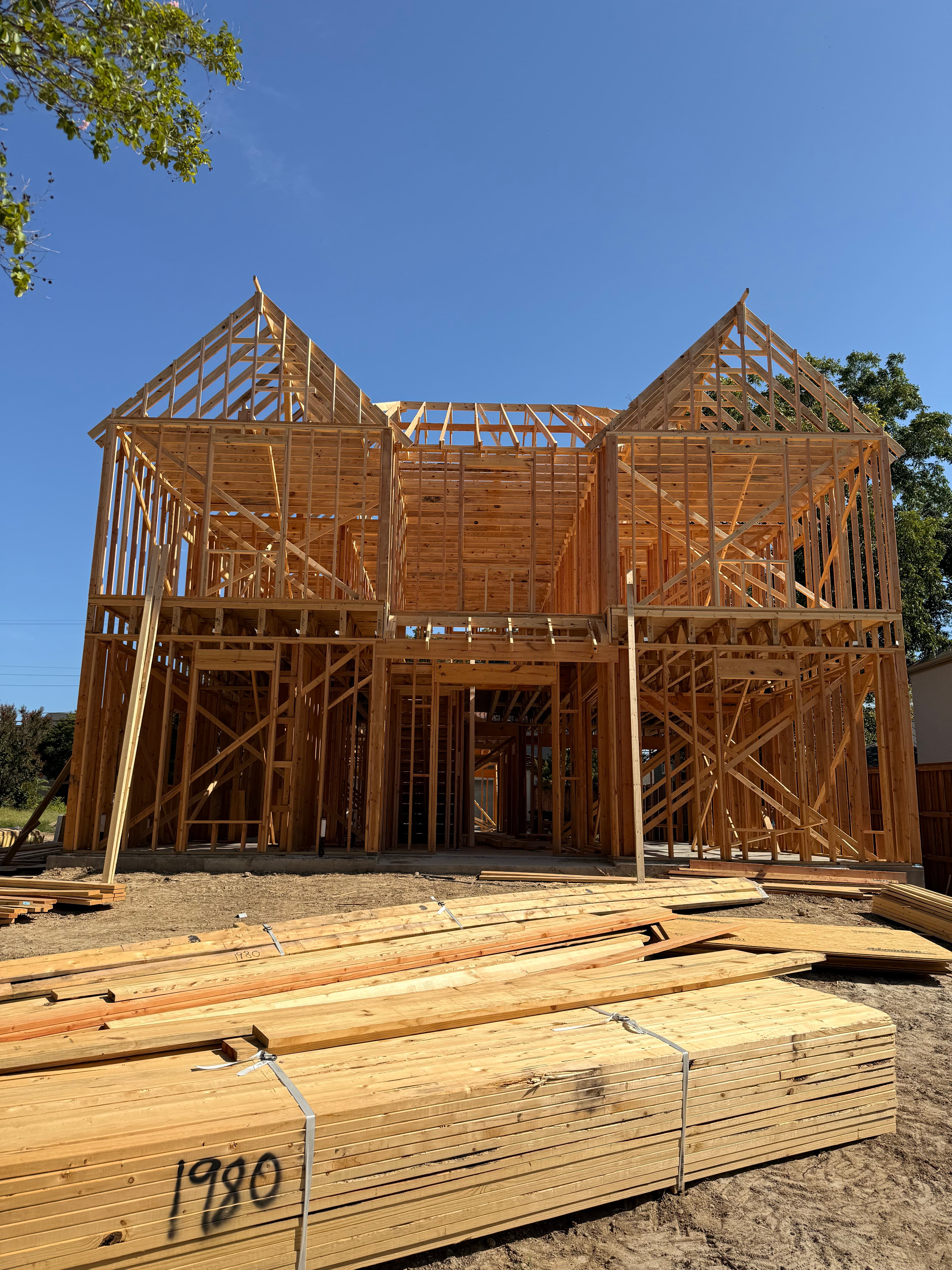 A two-story wooden house frame under construction with stacks of lumber in the foreground.